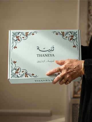 A hand holding a pale green Thaneya gift box decorated with painted dates and leaves
